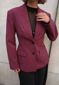 Bordo pritaikyta švarkas su aštriais apykaklėmis, dviem priekinėmis kišenėmis ir juodais mygtukais, dėvimas ant juodo raukšlėto topu, prieš neutralų foną.