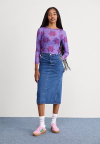Haut mauve à manches longues avec motif floral rose, associé à une jupe midi en denim bleu. Le modèle porte des baskets roses avec des rayures vertes et des chaussettes blanches.