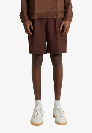 Uomo che indossa pantaloncini e camicia di lino marrone con calzini bianchi e scarpe da ginnastica bianche con suola in gomma, in piedi su sfondo uniforme.