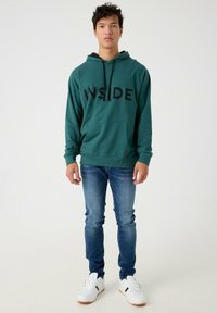 Sudadera color azul verdoso con el texto "INSIDE" en negro, bolsillo tipo canguro, cordón ajustable, llevada con jeans azules y zapatillas blancas.