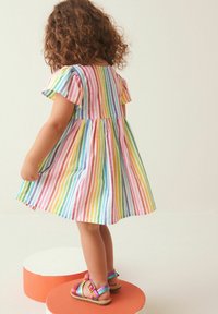 Mehrfarbig gestreiftes Kleid mit kurzen, flatternden Ärmeln, aus leichtem Stoff. Getragen mit bunten Riemchensandalen, das Kind mit dem Rücken zur Kamera.