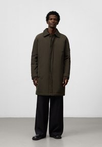 Cappotto imbottito lungo verde oliva con colletto, zip frontale e tasche laterali, abbinato a pantaloni neri larghi e scarpe scure.
