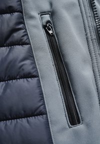 Podedna jakna v mornarsko modri in sivi barvi. Ima sivo tkaninsko sekcijo z žepom na zadrgo, kontrastne teksture in gladke odtise.