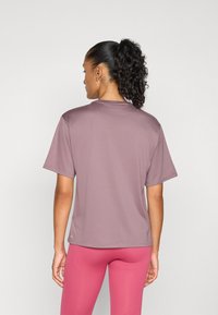 Femeie cu coada de cal bucle, îmbrăcată cu un tricou scurt cu mâneci scurte de culoare mov deschis și colanți roz ajustați, stând în fața unui fundal simplu de culoare deschisă.