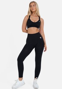 Schwarzer Sport-BH mit breiten Trägern und passenden schwarzen High-Waist-Leggings. Beide Teile bestehen aus dehnbarem, glattem Stoff. Weiße Logodetails.