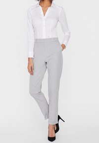 Chemise blanche à manches longues avec boutons, associée à un pantalon gris clair ajusté et des talons hauts noirs. Tissu lisse avec une texture subtile.