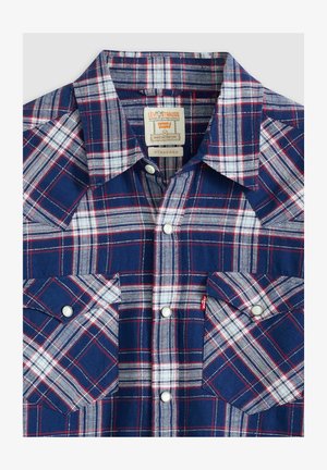 Chemise à carreaux bleu marine, blanche et rouge. Présente un col pointu, deux poches poitrine et des boutons-pression argentés. Tissu texturé.