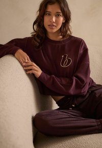 Burgundské pletené tričko s logem zlatého písmene "b", spárované s odpovídajícími kalhotami. Hladká textura, volný střih a žebrované manžety.