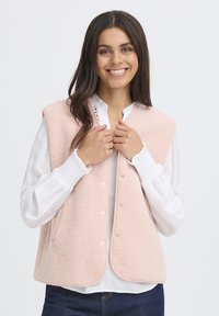 Rózsaszín, ujjatlan mellény, puha, texturált bolyhos anyagból, kerek szegéllyel, patentgombos elejével és kontrasztos díszítéssel, fehér blúzzal párosítva.