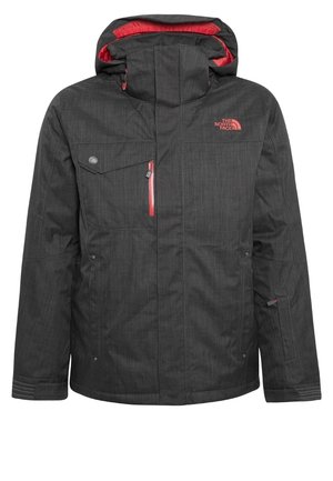 Muž nosící černé a červené lyžařské boty, tmavě šedé kalhoty s kapsami, černou bundu se červeným zipem a hnědý svetr, stojící na jednobarevném pozadí.