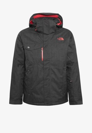 Homme portant des chaussures de ski noires et rouges, un pantalon cargo gris foncé, une veste noire avec une fermeture éclair rouge, et un pull marron, debout devant un fond uni.