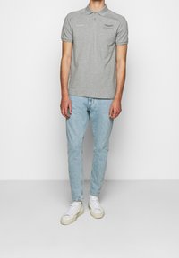 Šedé polo tričko s krátkými rukávy, se vyšívaným textem a logem, spárované s světlomodrými džíny a bílými teniskami.