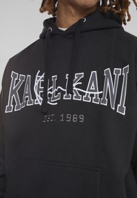 Svart hoodie med vit broderad text "KALIKANI" och året "EST. 1989." Fabrik i bomullsblandning, dragsko i huvan och känguruficka.