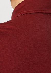 Gros plan d'un col de chemise en tissu rouge et du haut du dos d'une personne à la peau claire sur un fond uni.