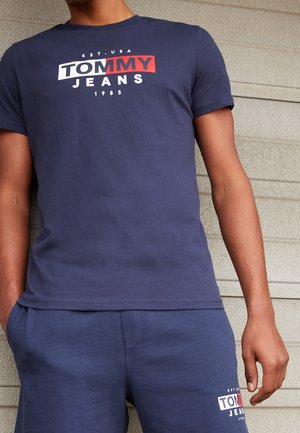 Mężczyzna w granatowej koszulce Tommy Jeans i dopasowanych szortach z białym i czerwonym logo, stoi przy teksturowanej ścianie.