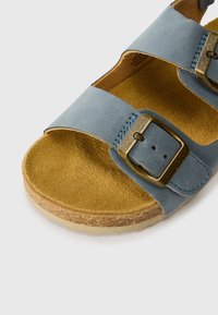 Μπλε δερμάτινη σαγιονάρα με δύο ρυθμιζόμενους ιμάντες, μπρούντζινες αγκράφες, φιάλη από φελλό και ταμπά σουέτ υφή. Ελαφρύς σχεδιασμός και επίπεδη σόλα.