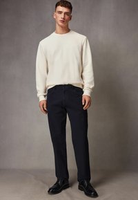 Cream sweatshirt with textured fabric, paired with dark navy trousers. Model wearing black lace-up shoes, standing against a gray background.