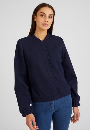 Veste en tissu doux bleu marine avec un col rond, des poignets élastiques et une fermeture frontale simple ; elle présente un design cropped et deux poches latérales.