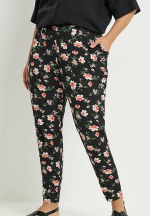 Vrouw draagt zwarte, nauwsluitende broek met roze en witte bloemenprint, gecombineerd met een zwart topje en zwarte sandalen.