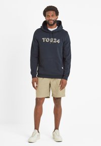 Man wearing navy TOG24 hoodie, beige shorts, and white sneakers standing against a plain white background.