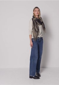 Écharpe à motifs gris et noir avec franges, drapée sur un pull léger, associée à un jean large bleu et des bottines noires.