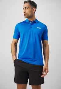 Blå polo-shirt med knapkrave, udstyret med et hvidt logo på brystet og korte ærmer, sammen med sorte shorts.