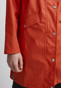 Giacca impermeabile rossa con bottoni a pressione, due tasche frontali e una texture liscia. Presenta una mano che tiene il tessuto vicino all'orlo.