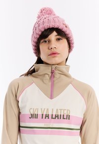 Beige sporttop met een kwart-rits en roze details, voorzien van de tekst "SKI YA LATER". Roze gebreide beanie met een pompon bovenop.