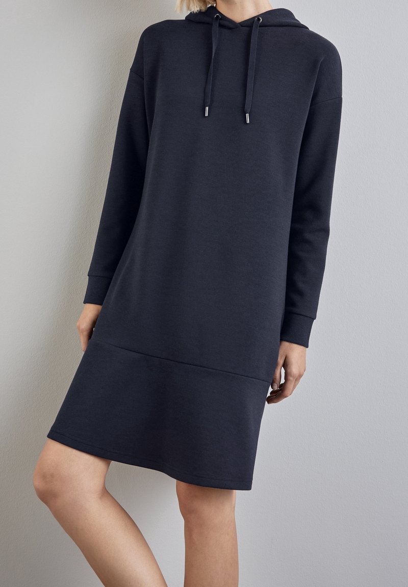 Femme portant une robe à capuche noire à manches longues, debout contre un mur uni de couleur claire.