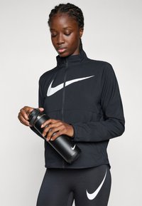 Schwarze leichte Zip-Jacke mit hohem Kragen, die ein großes weißes Nike-Swoosh-Logo zeigt. Das Modell hält eine schwarze Wasserflasche mit einem Deckel.