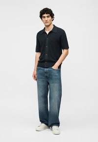 Νέος άνδρας στέκεται με το ένα χέρι στην τσέπη, φοράει μαύρο κοντομάνικο πουκάμισο με κουμπιά, φαρδιά μπλε τζιν και άσπρα σνίκερς.