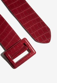 Rode riem van nep-krokodillenleer met een glanzende, vierkant gesneden gesp en een gestructureerd oppervlak met een rechthoekige opening voor de pen van de gesp.