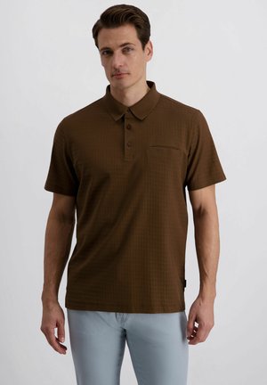 Homme portant un polo marron à manches courtes et un pantalon gris clair, debout devant un fond blanc uni.