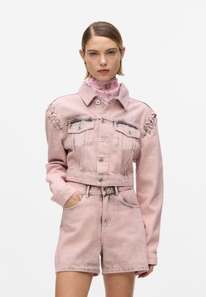Žena v svetloružovej džínsovej skrátenej bunde s viazaním na ramenách a zodpovedajúcich vysokých šortkách, stojaca pred bielym pozadím.