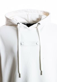Sudadera blanca con capucha, hecha de suave algodón, que presenta una superficie texturizada, cordones ajustables con puntas de metal y una etiqueta en el frente.