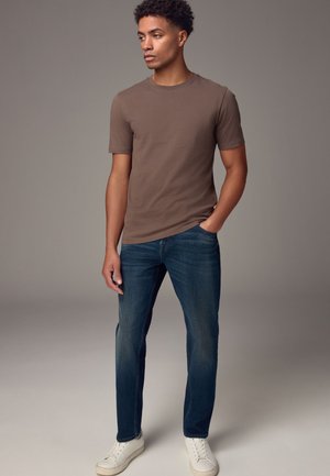 Junger Mann trägt braunes kurzärmeliges T-Shirt, blaue Jeans und weiße Sneaker, steht mit einer Hand in der Tasche vor einfachem Hintergrund.