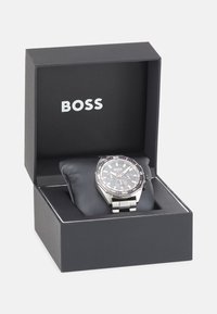 Reloj analógico negro con correa de acero inoxidable plateado, exhibido sobre una suave almohadilla negra dentro de una caja negra etiquetada "BOSS".
