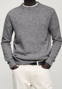 Personne à la peau foncée portant un pull en maille gris moucheté par-dessus une chemise blanche rentrée dans un pantalon crème avec une ceinture noire, les mains dans les poches.