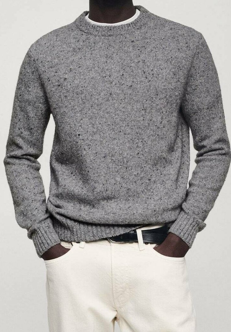 Personne à la peau foncée portant un pull en maille gris moucheté par-dessus une chemise blanche rentrée dans un pantalon crème avec une ceinture noire, les mains dans les poches.