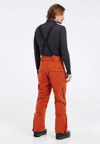 Orange vattentäta skidbyxor med justerbara svarta hängslen, sidoknappar och förstärkt sömnad, tillsammans med en långärmad svart topp.