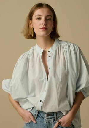 Jonge vrouw draagt een oversized wit blouse met knopen, blauwe spijkerbroek, gouden oorbellen en ring, staand tegen een beige achtergrond met handen in de zakken.