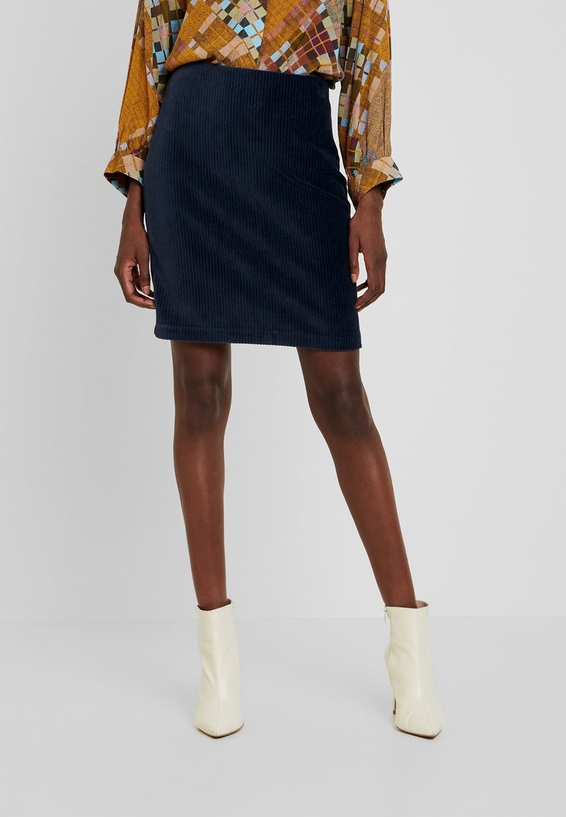 Femme vêtue d'une jupe en velours côtelé bleu foncé, d'un chemisier à motifs multicolores et de bottines blanches, debout devant un fond uni.