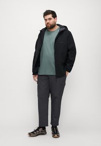 Black lightweight jacket, green t-shirt, and grey cargo pants. Wearing patterned trail shoes. Outdoor apparel suitable for varied conditions.
