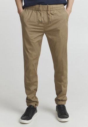Men's khaki jogger pants with an elastic waistband, adjustable drawstring, and cuffed hems. Paired with black sneakers.