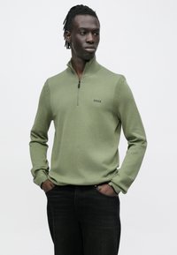 Zielony sweter z półgolfem na zamek, wykończony prążkowanymi elementami, wykonany z miękkiego materiału. Posiada subtelne logo na piersi i jest zestawiony z ciemnymi jeansami.