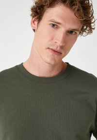 Grøn bomulds t-shirt med rund hals og tekstureret finish, med ribbede detaljer ved halsåbningen og korte ærmer. Nærbillede af stoffet.