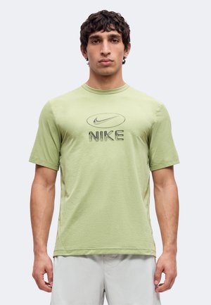 Νέος άνδρας που φοράει ένα ανοιχτό πράσινο T-shirt Nike με κοντά μανίκια και λευκά σορτς, στέκεται μπροστά σε ένα απλό φωτεινό φόντο.