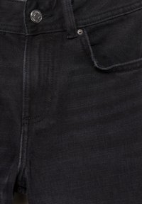 Gros plan sur un jean en denim noir montrant la poche avant, les passants de ceinture, les coutures et un bouton en métal avec texte gravé.