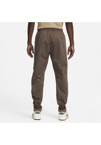 Muž oblečený v hnědých cargo kalhotách a bílých teniskách, stojící zády k fotoaparátu na jednobarevném bílém pozadí.