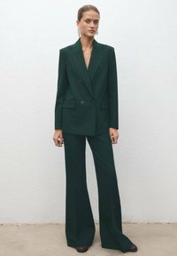 Tamnozeleni sako s dva reda gumba i uvučenim reverima, uparen s širokim hlačama iste boje. Glatka tkanina i minimalistički dizajn.